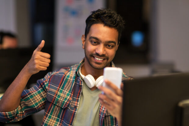 technology, communication and deadline concept - creative man with smartphone having video call at night office and showing thumbs up