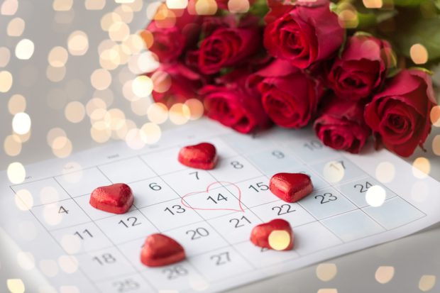 Calendar with a heart drawn around February 14th, surrounded by red chocolate hearts and a bunch of red roses