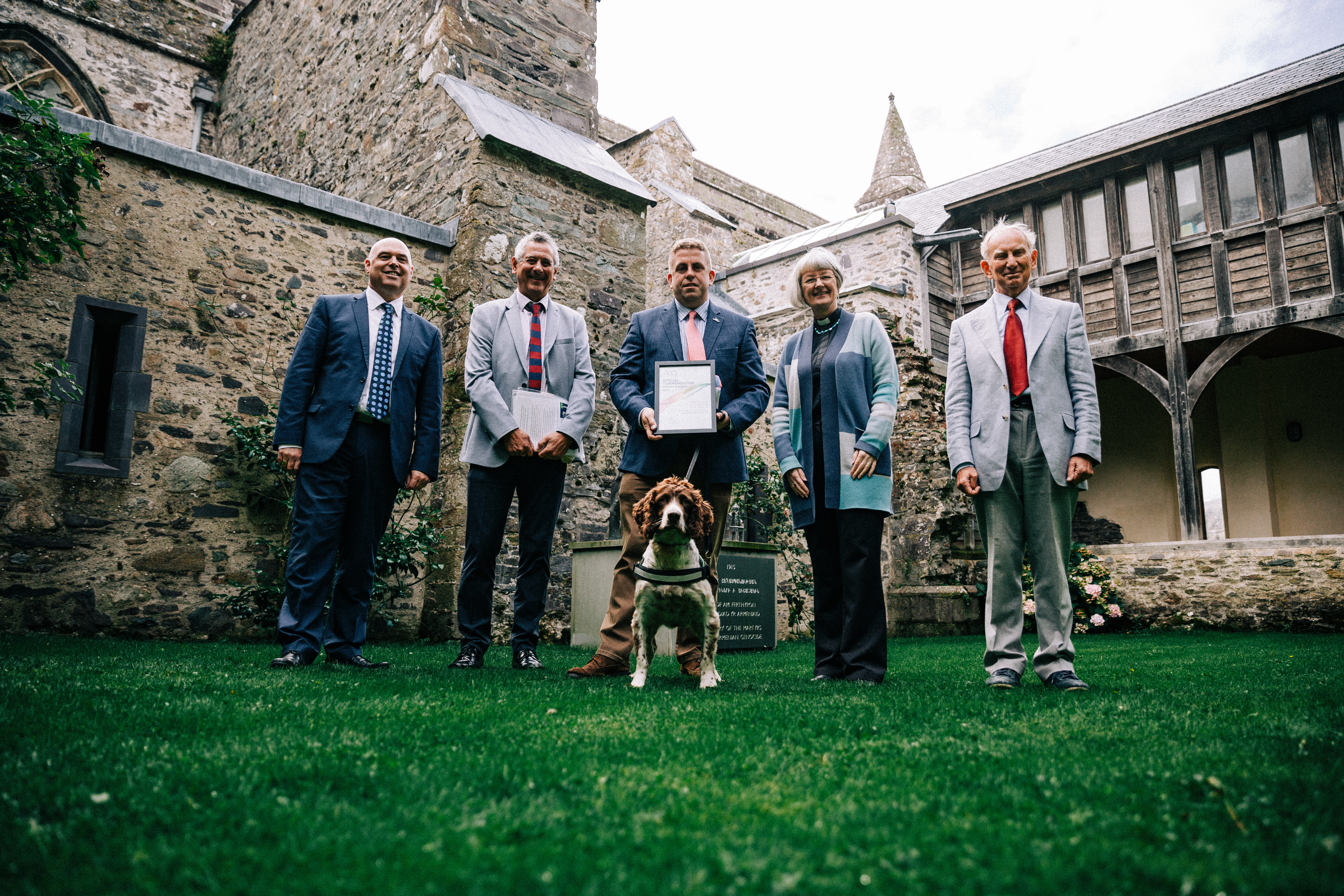 Five representatives gather in front of a cathedral with Scamp the sniffer dog holding certificates.