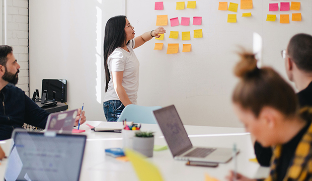 People sitting around a desk working and chatting whilst a woman in the background is pointing to post it notes on a whiteboard