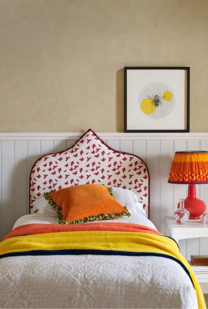 A bed with orange cushion and yellow throw, a patterned headboard, picture of a bee on the wall and bright orange coloured lamp on bedside table