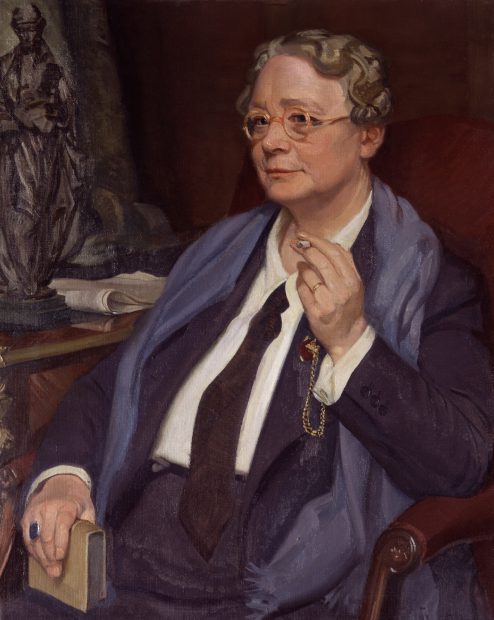 Dorothy L Sayers sat in a chair smoking a cigarette, wearing a suit and tie with scarf