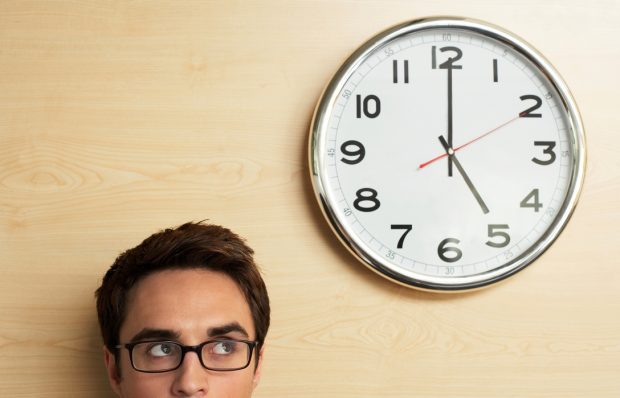 Man wearing glasses glancing up at a clock on the wall showing 5pm