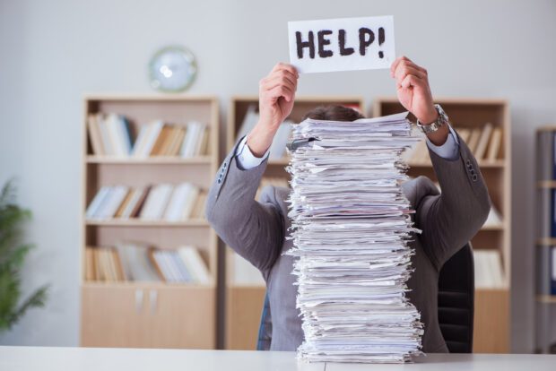 Image of business man hidden behind a large stack of paperwork holding sign that says help