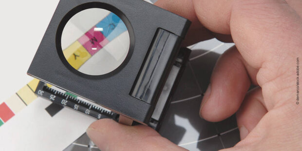 Hand holding a magnifying loupe to closely inspect color swatches on a printed sheet.